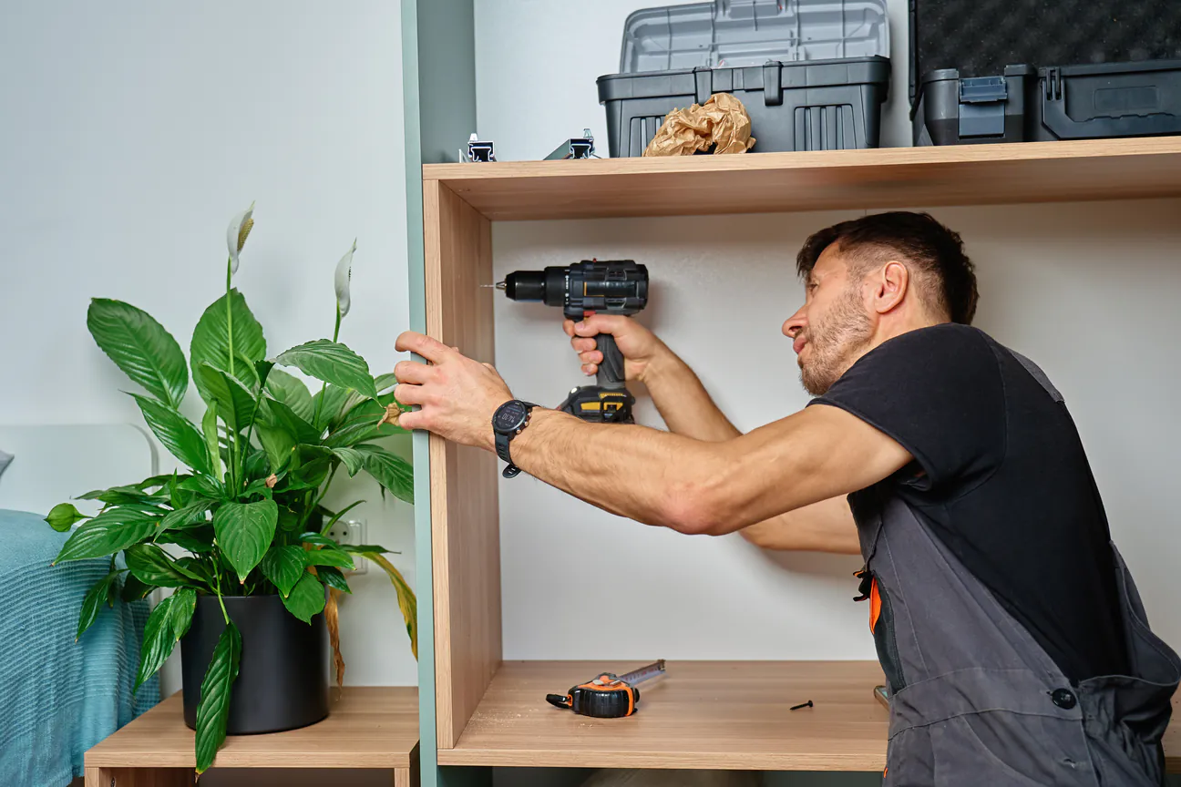 A man driving a screw into a wall shelf with a cordless drill.