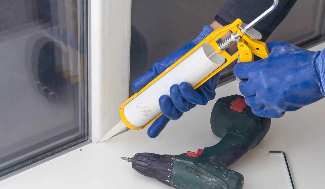 Gloved hands running a caulk gun along the edge of a white frame