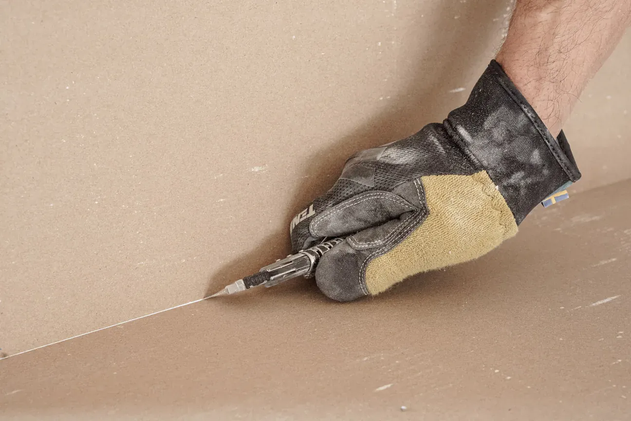 A gloved hand scoring a sheet of drywall with a utility knife