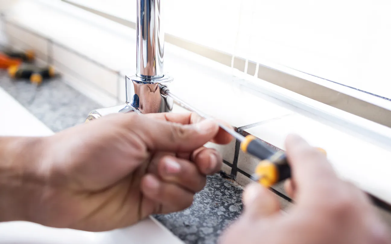 Two hands steadying a chrome faucet while a screwdriver turns a fixing at its base