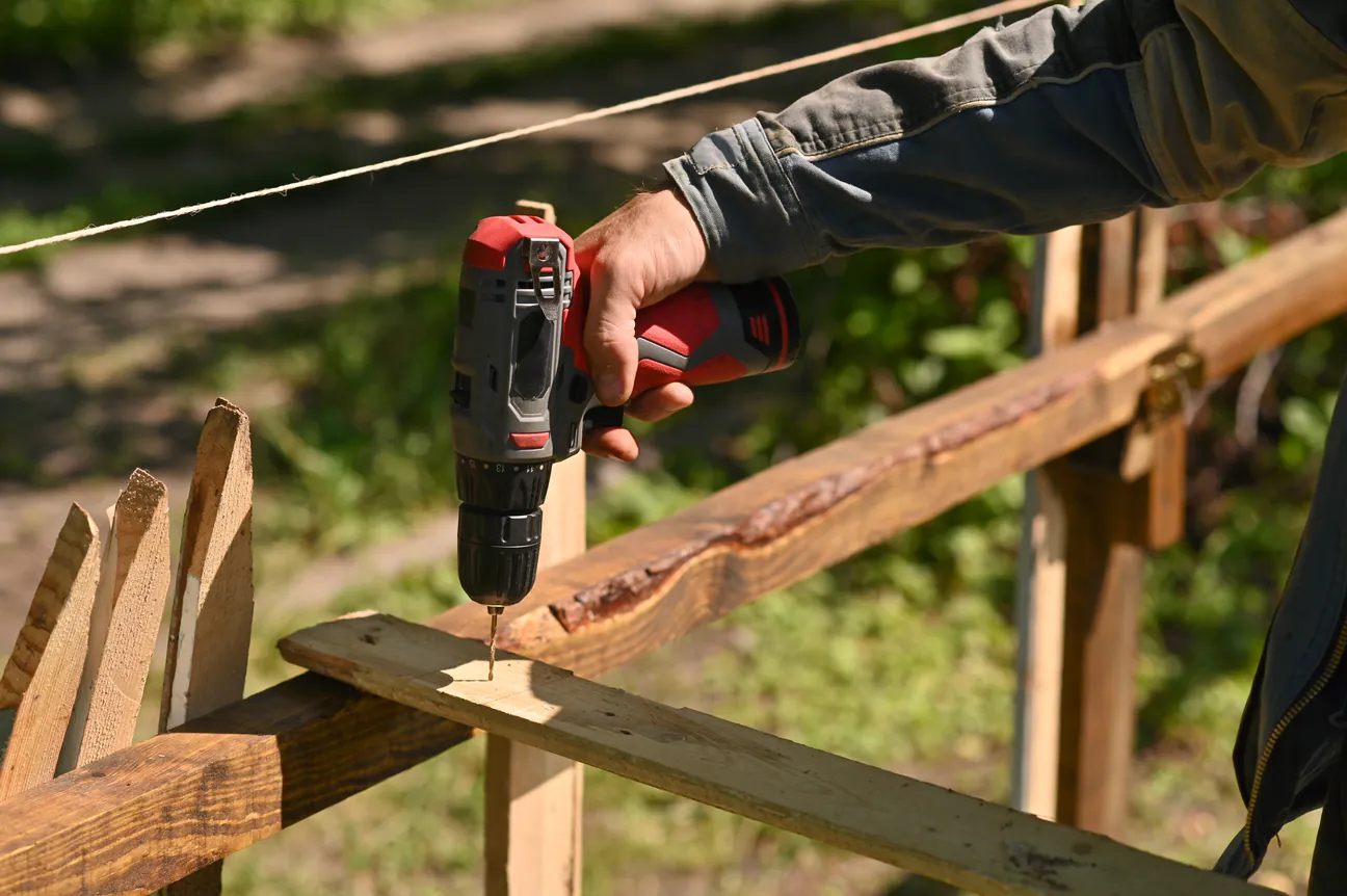 A cordless drill driving a screw into a wooden fence rail with a string line stretched along the fence