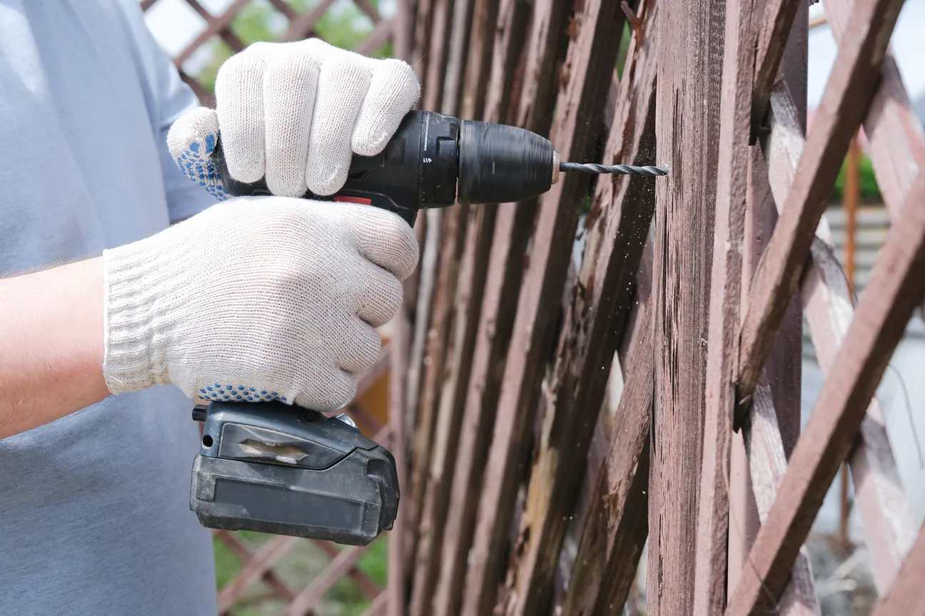 Gloved hands driving a drill into a weathered wood fence