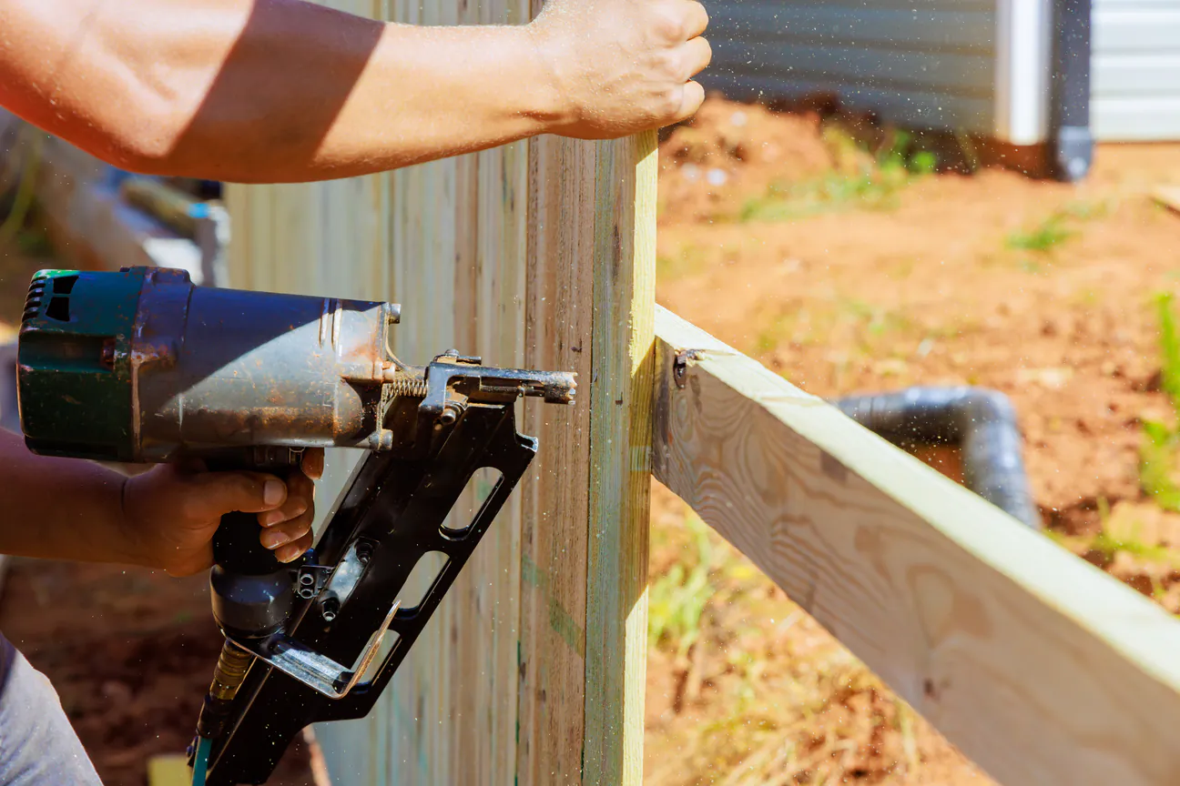 A framing nailer fastening a wooden fence rail to a post