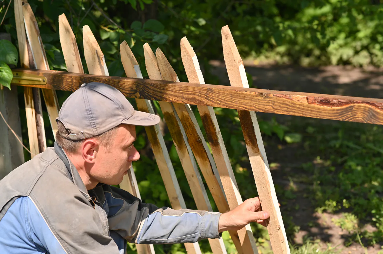 A worker fitting a fresh picket into a weathered wooden fence outdoors