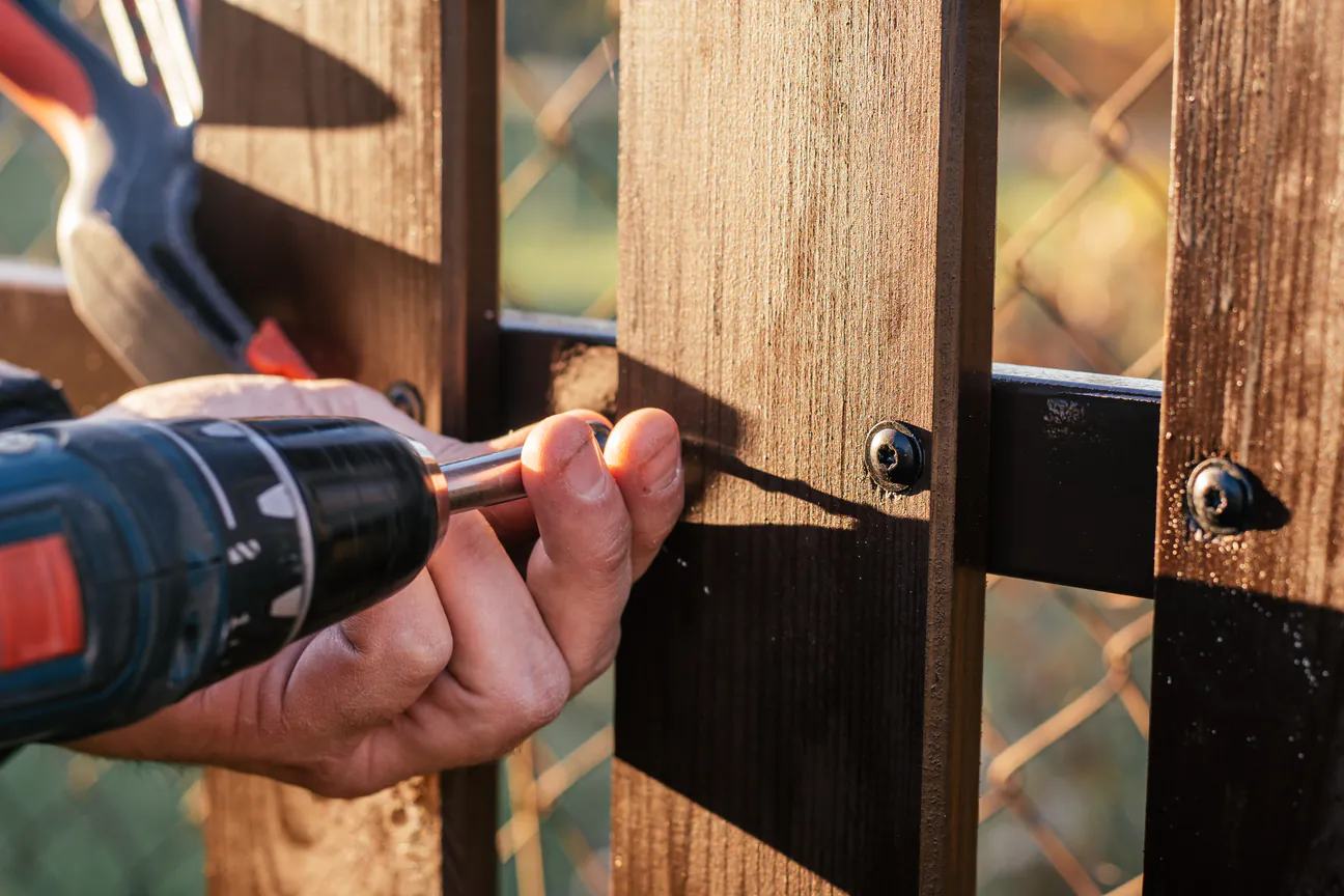 A cordless drill driving screws through a fence board into the rail