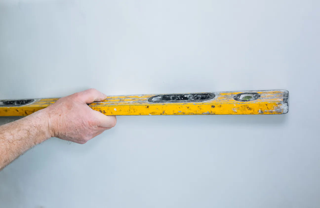 A hand holding a yellow spirit level flat against a white wall