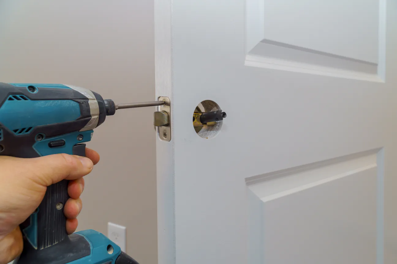 A cordless driver fastening a latch plate to the edge of a white interior door