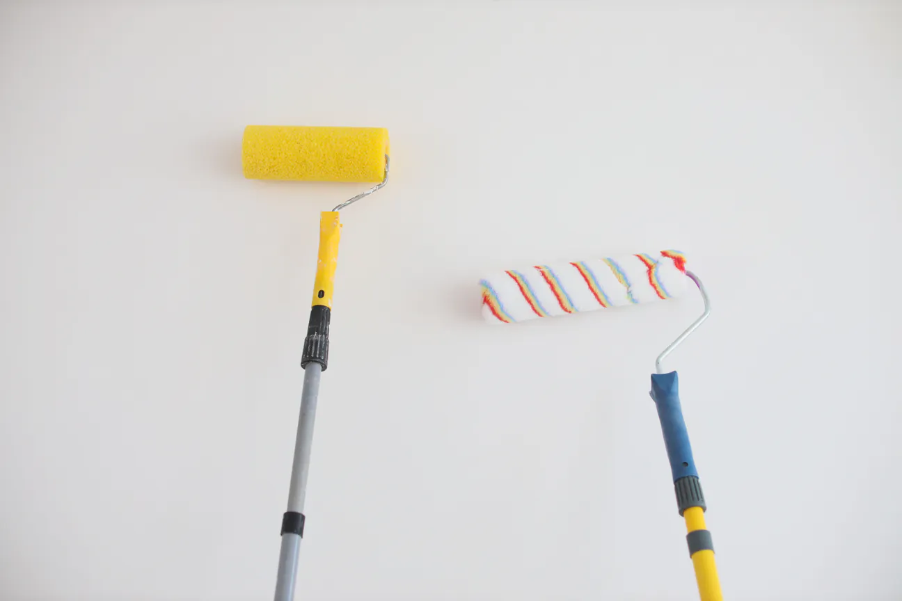 Two paint rollers on extension poles resting against a white wall