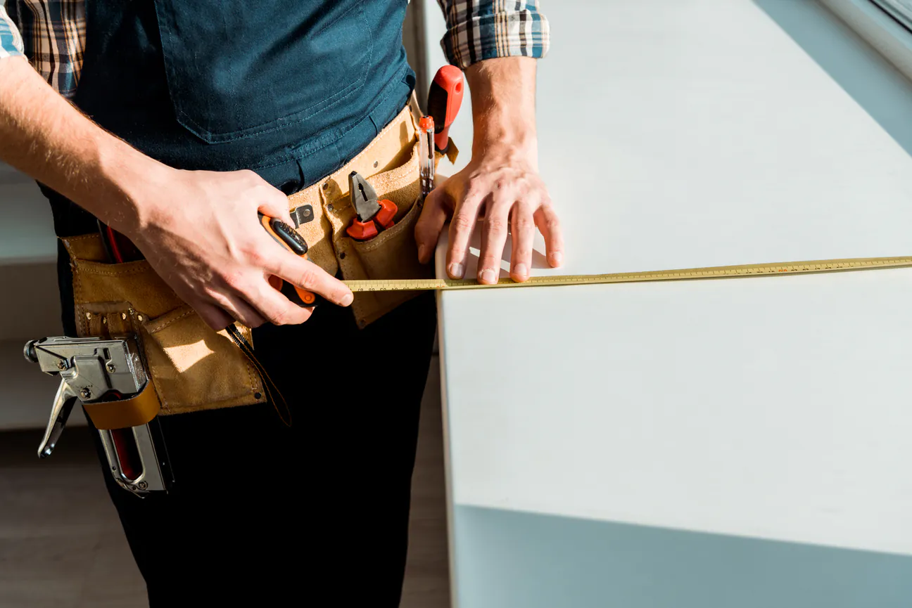 An installer wearing a tool belt running a tape measure along a white windowsill