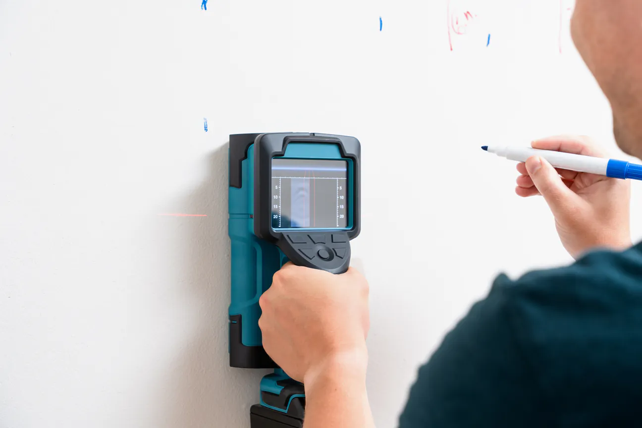 A wall scanner held against a wall while a hand marks the stud line