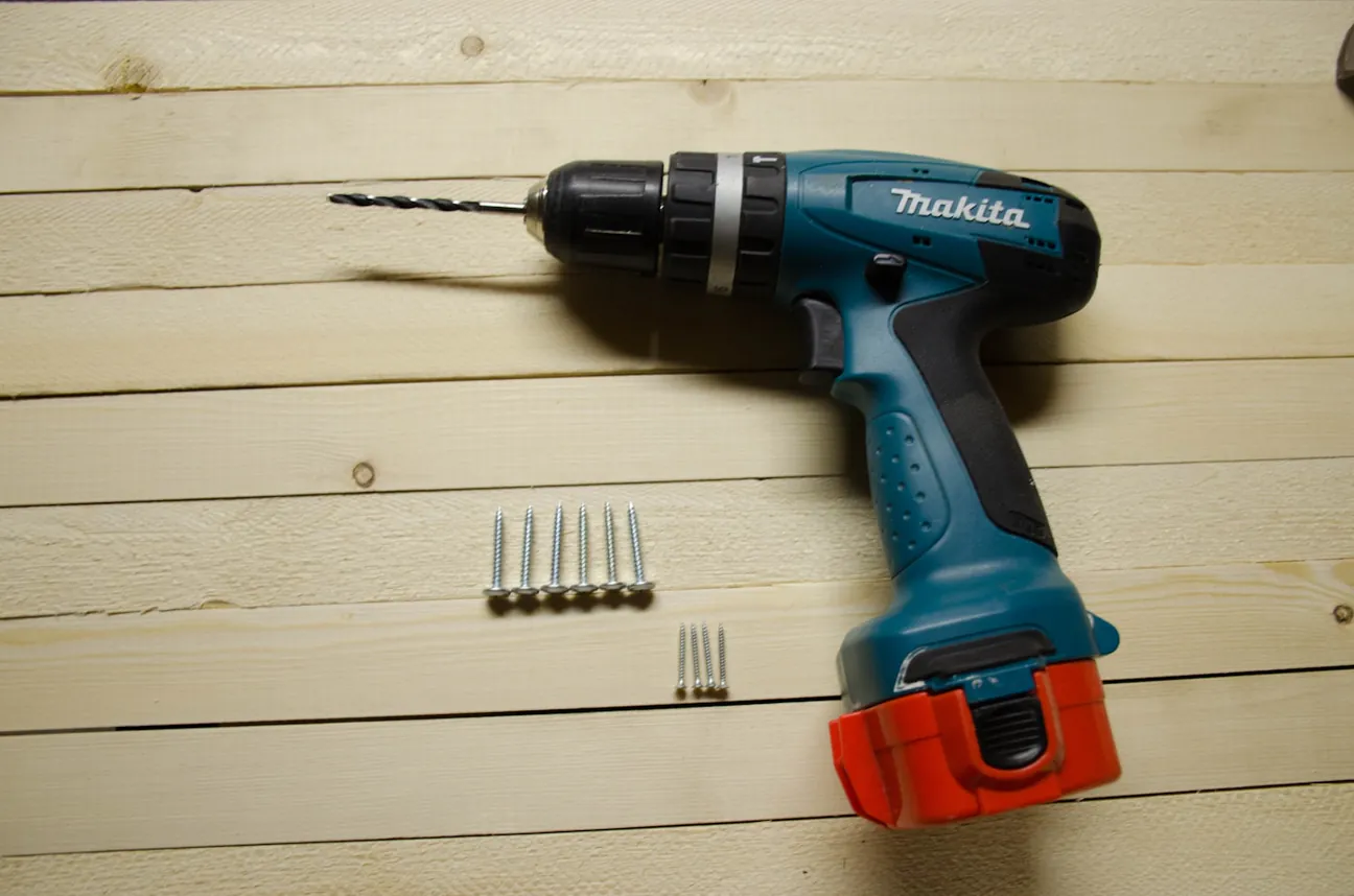 A cordless drill and rows of screws laid out on a wood surface