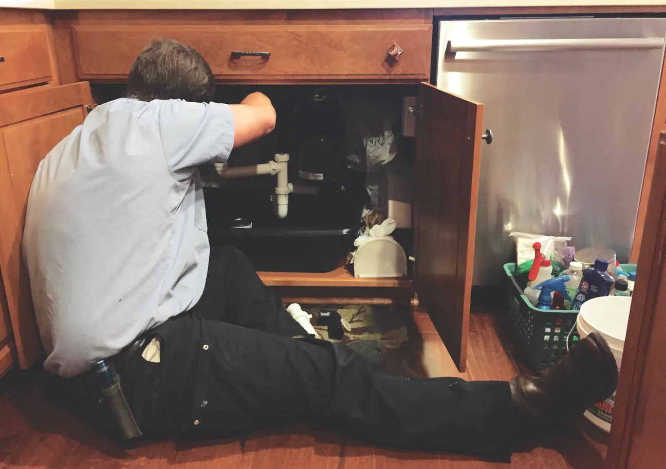 A worker sitting at an open kitchen cabinet working on the pipes under the sink